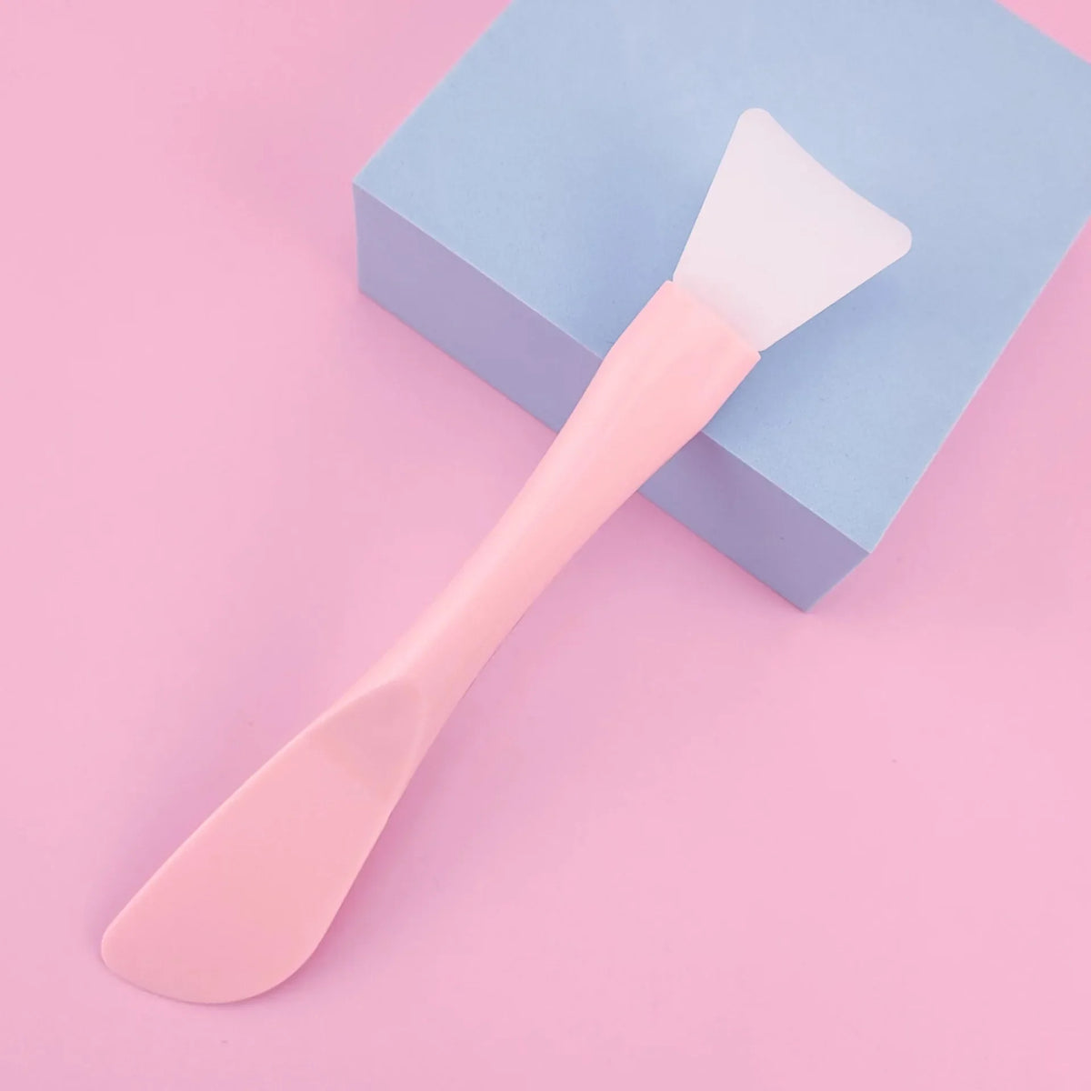 A pink silicone facial mask brush with an integrated scraper rests on a light blue square box.