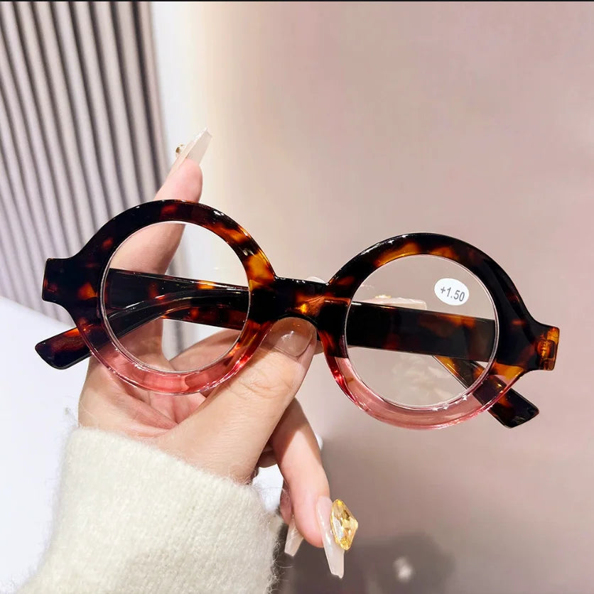 A hand holds a pair of round, tortoiseshell-patterned reading glasses with pink acetate frames.
