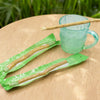 Une tasse en verre et des cure-dents miswak naturels enveloppés dans du matériel vert sont présentés sur une surface en bois.