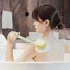 A woman is using a long-handle loofah back scrubber with a soft mesh sponge while bathing.