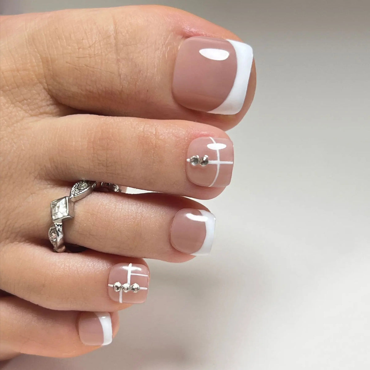 Eine Hand zeigt eine Nageldesign mit sanfter Nude-Basis, weißen French-Tips und zarten silbernen Stud-Akzenten.