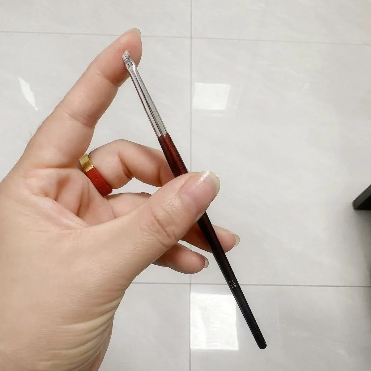 Une main tient un pinceau de maquillage à fondation anglé avec un manche foncé et une pointe argentée sur un fond blanc.