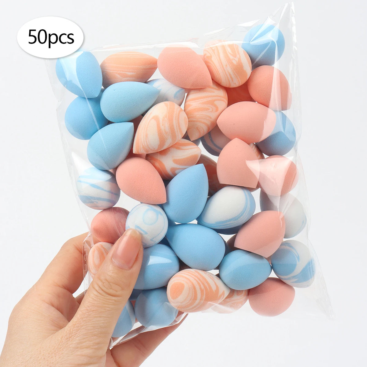 A hand holds a clear plastic bag filled with 50 reusable expandable makeup sponges in various pastel shades of blue and pink.