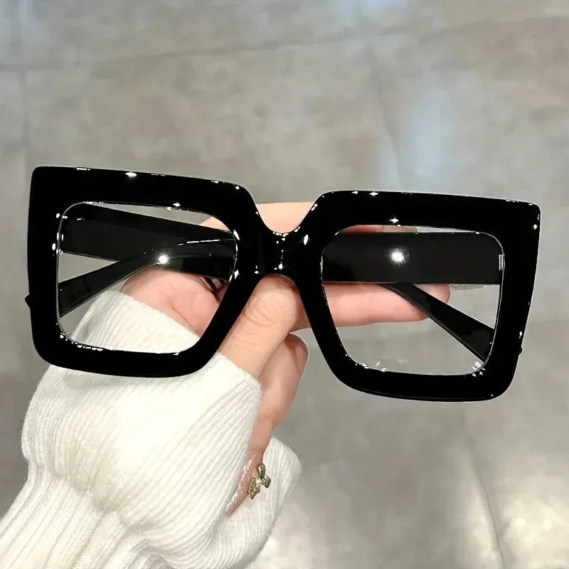 A hand holds a pair of black square eyeglasses with clear lenses against a light background.