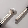 A silver and black double-ended eye shadow brush with a dense synthetic hair tip is shown on a gray background.