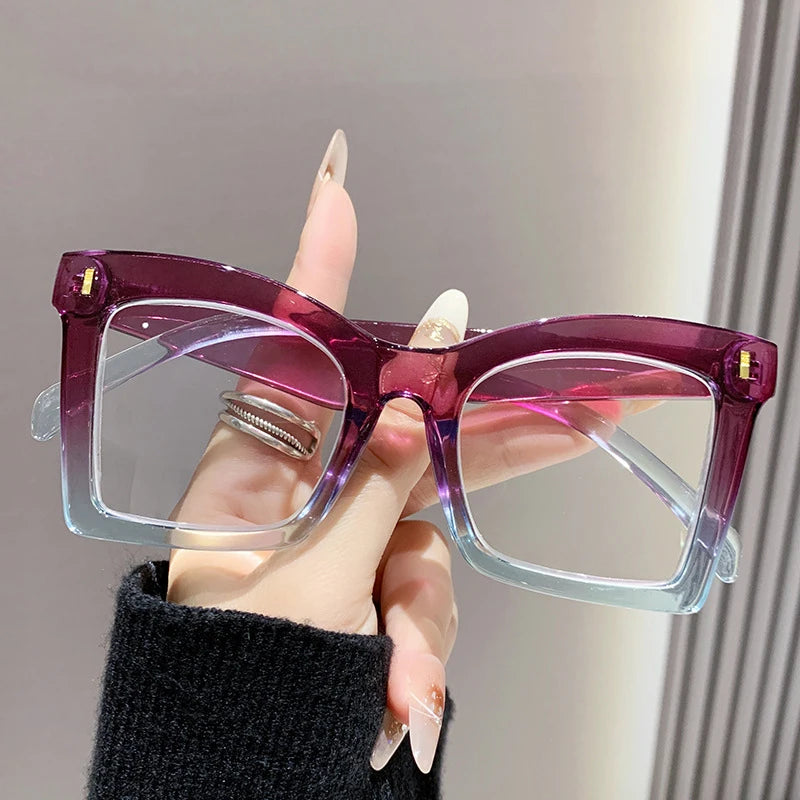 A hand holds a pair of purple square frame reading glasses with clear lenses against a light background.