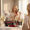 A woman holds a red hair styling tool and dryer in a bright bedroom setting.