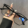 A hand holds a pair of black minimalist square reading glasses with gold accents against a blurred background.