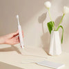 A hand holds a white sonic electric toothbrush and its charging cord next to a white charging base.