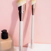 A set of two natural-bristled makeup brushes with wooden handles displayed against a pink background.