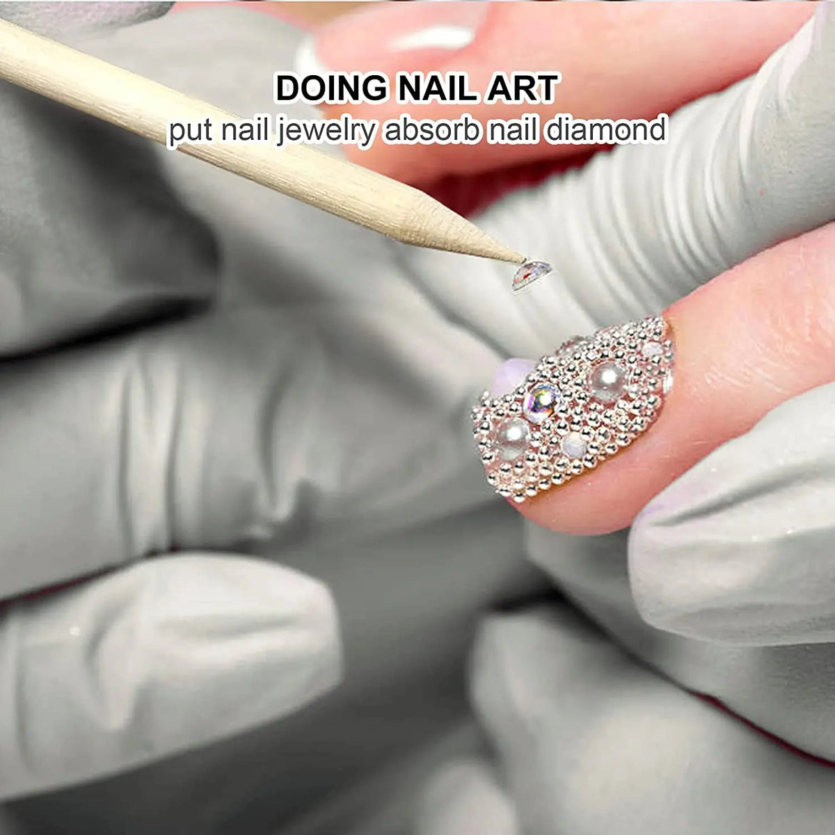 A hand wearing a glove is using a wooden stick to apply nail jewelry onto a fingernail for nail art.