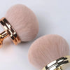 A large, round makeup brush with light beige bristles and a gold-toned handle is shown against a white background.