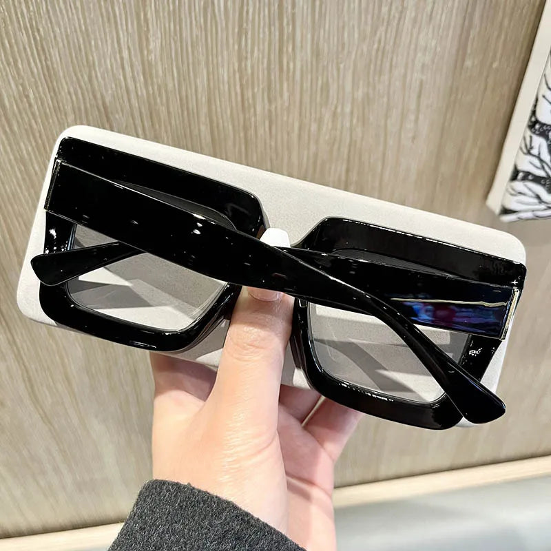 A hand holds a pair of black square eyeglasses with clear lenses against a light background.