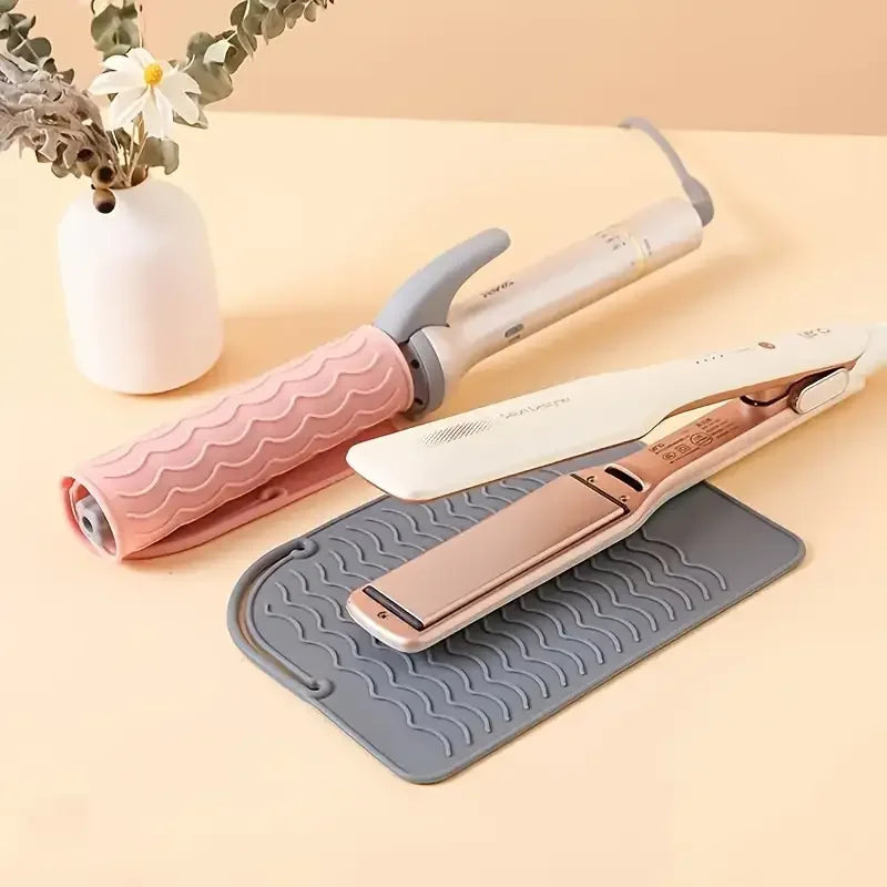 A silicone heat resistant mat is placed under a hair styling tool, with a white vase and some greenery in the background.