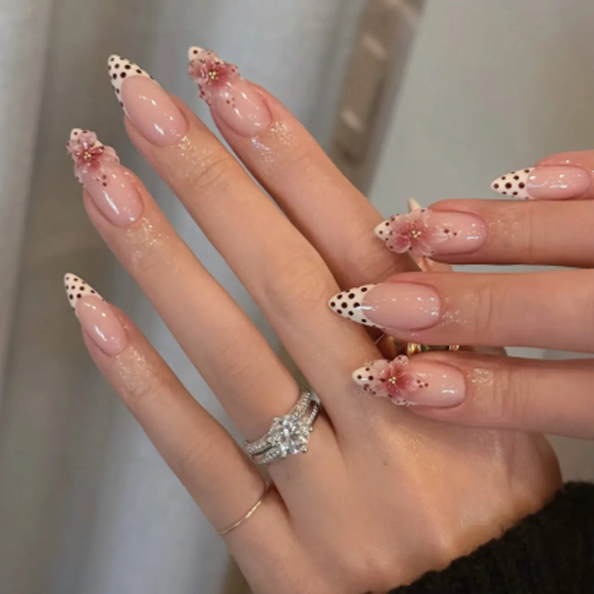 Gros plan d'une main présentant des kits de vernis press-on en forme de fleur d'amande avec des accents rose pâle et or.