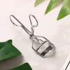 A silver professional eyelash curler tool resting on a white surface with green leaves.