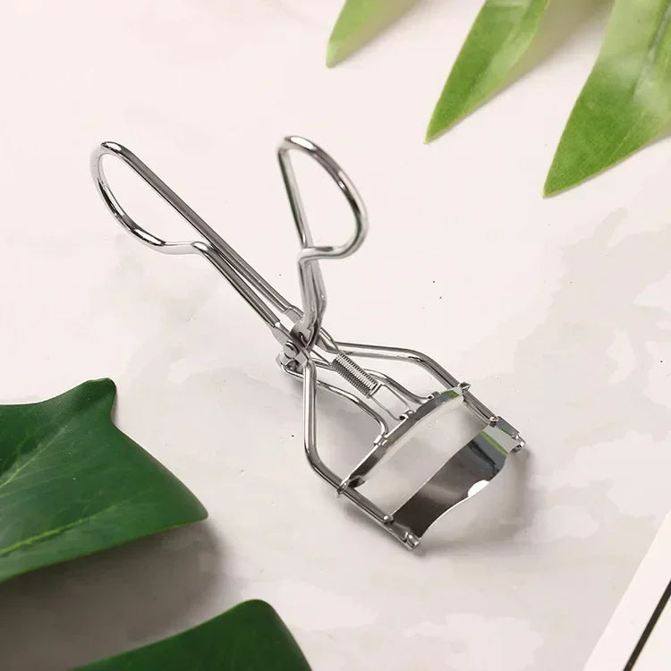 A silver professional eyelash curler tool resting on a white surface with green leaves.