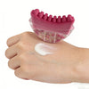 A hand holds a pink natural rubber gua sha tool with pink bristles and a white massage cream on the skin.