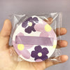 A white makeup puff set with purple and yellow floral designs is shown inside a clear plastic storage bag.