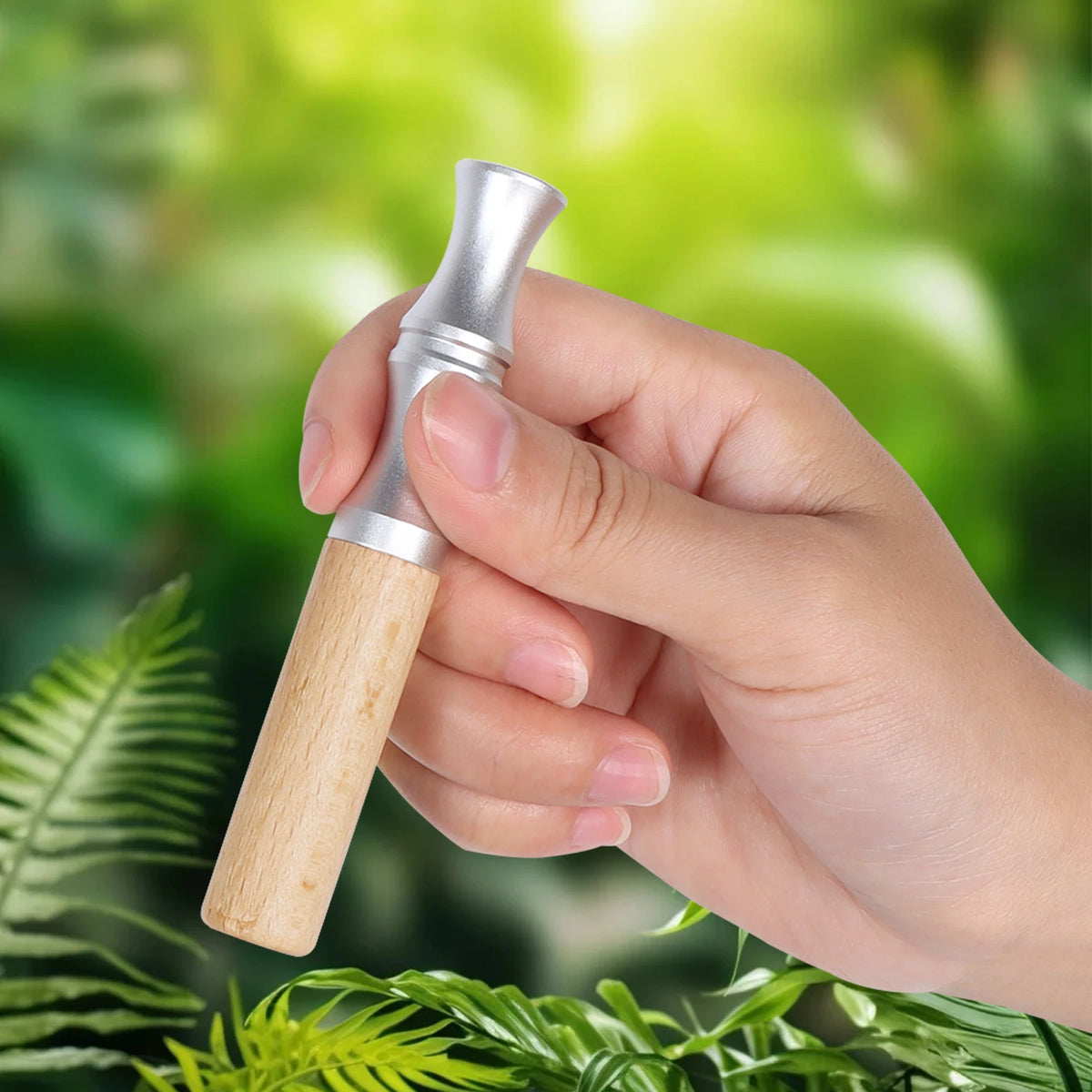 A hand holds a natural wooden stop smoking stick diffuser with a silver cap against a blurred green background of foliage.