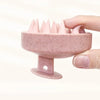 A hand holds a pink silicone scalp massage and body brush with numerous pointed bristles.