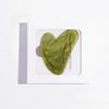 A smooth, jade-green Gua Sha tool shaped like a heart is shown on a white background.