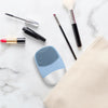 A sonic facial cleansing brush and various beauty tools are arranged on a white marble surface next to a beige towel.