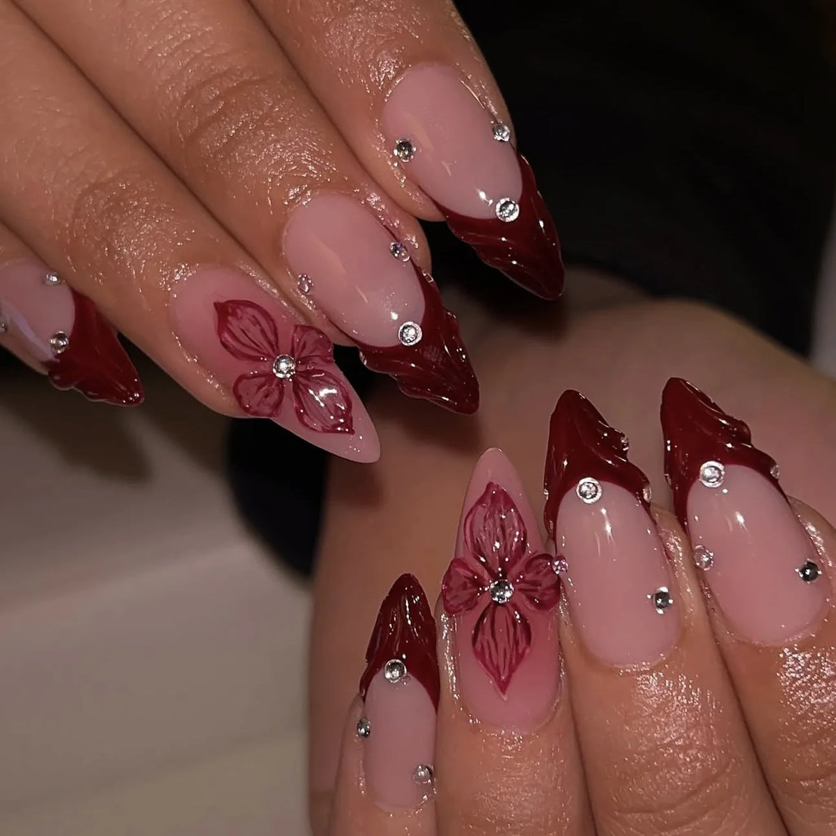 Gros plan d'une main montrant des ongles press-on français roses aux amandes avec de petits strass et des accents floraux.