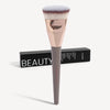 A professional flat kabuki foundation brush with a pink ferrule and dark handle resting on a black box.