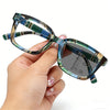 A hand holds a pair of blue and green photochromic reading glasses with pink accents.