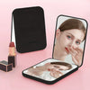 A compact LED makeup mirror with a foldable frame is shown in black and pink tones, displaying a magnified view of a woman's 