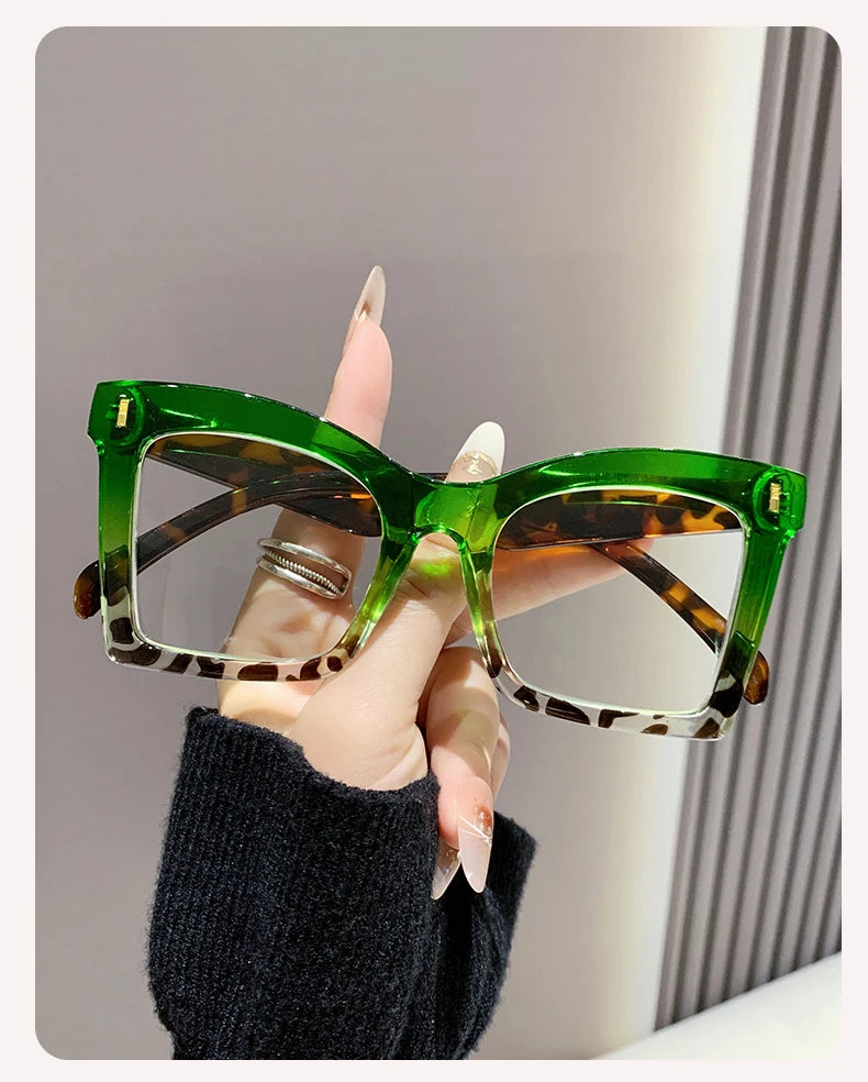 A hand holds a pair of green-framed reading glasses with tortoiseshell lenses against a light background.