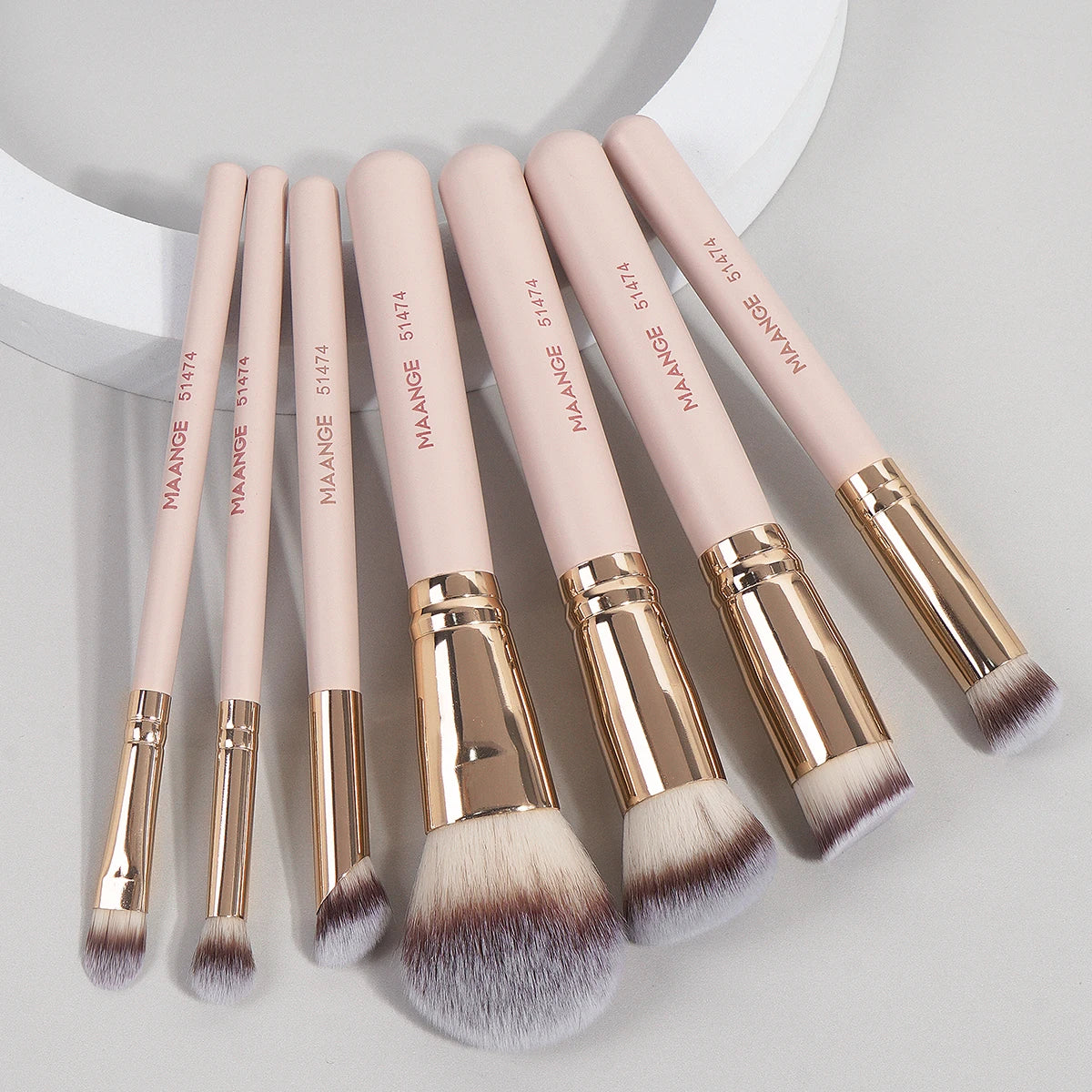 A set of seven pink makeup brushes with gold ferrules and fluffy synthetic bristles arranged diagonally on a light gray surfa