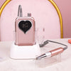A pink cordless rechargeable nail drill with a heart-shaped body and a drill bit resting on a white stand.