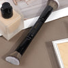 A black double-ended makeup brush with a silver handle and bristles rests on a light-colored box.