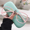 A hand holds a pair of pink-framed reading glasses with gold accents resting on a mint green silicone case.