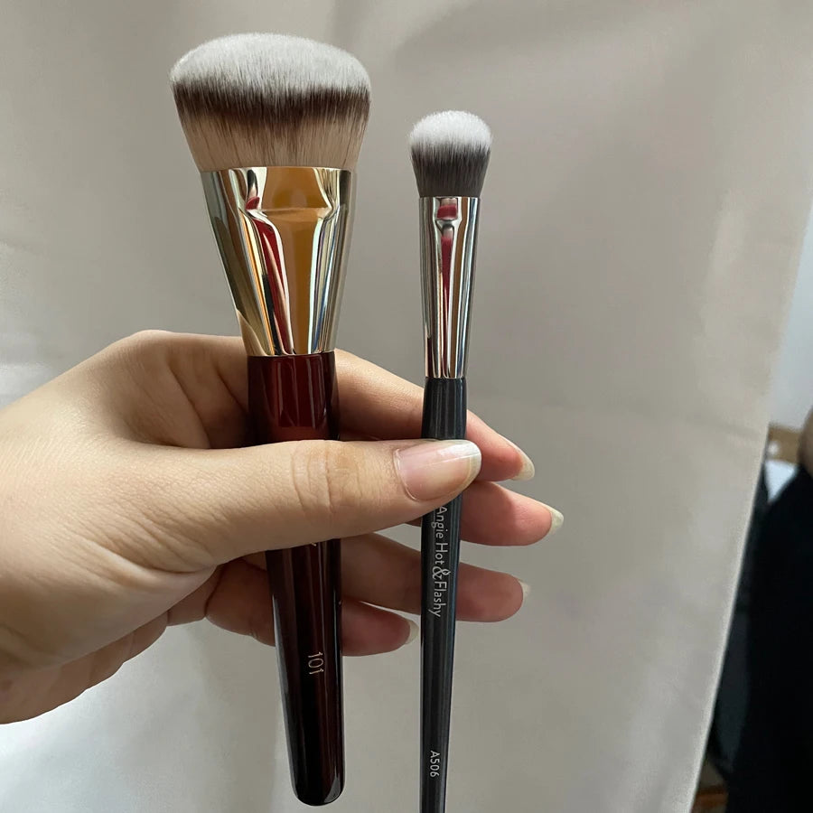 Une main tient un pinceau de maquillage liquide angulé avec une ferule dorée et une poignée noire.