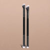 A pair of black makeup brushes with silver bristles displayed against a beige background.