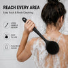 A woman is using a black long handle silicone body brush to clean her back with soap foam.