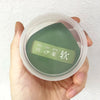 A hand holds a clear storage box containing an air cushion makeup puff set with a green pad inside.