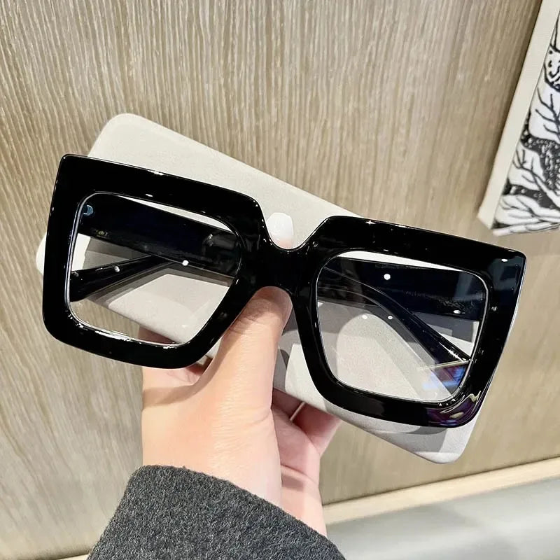 A hand holds a pair of black square eyeglasses with clear lenses against a light background.