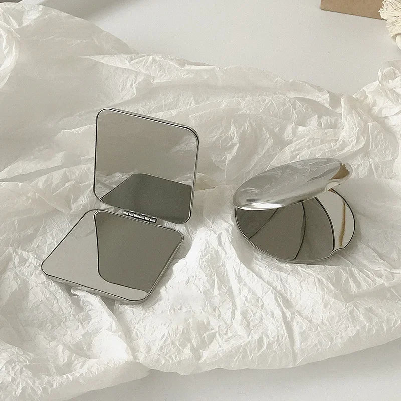 A stainless steel folding mirror is shown open and closed on crumpled white tissue paper.