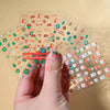 A hand holds a sheet of Christmas Santa Claus 3D nail art stickers with glitter designs showing various holiday patterns.