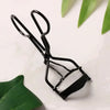 Black professional eyelash curler tool resting on a white surface with green leaves.