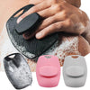 A hand is holding a black silicone body scrubber, with other variations of the exfoliating tool displayed nearby.