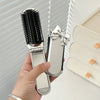 A hand holds a silver portable compact comb with an attached mirror and a black bristled comb.