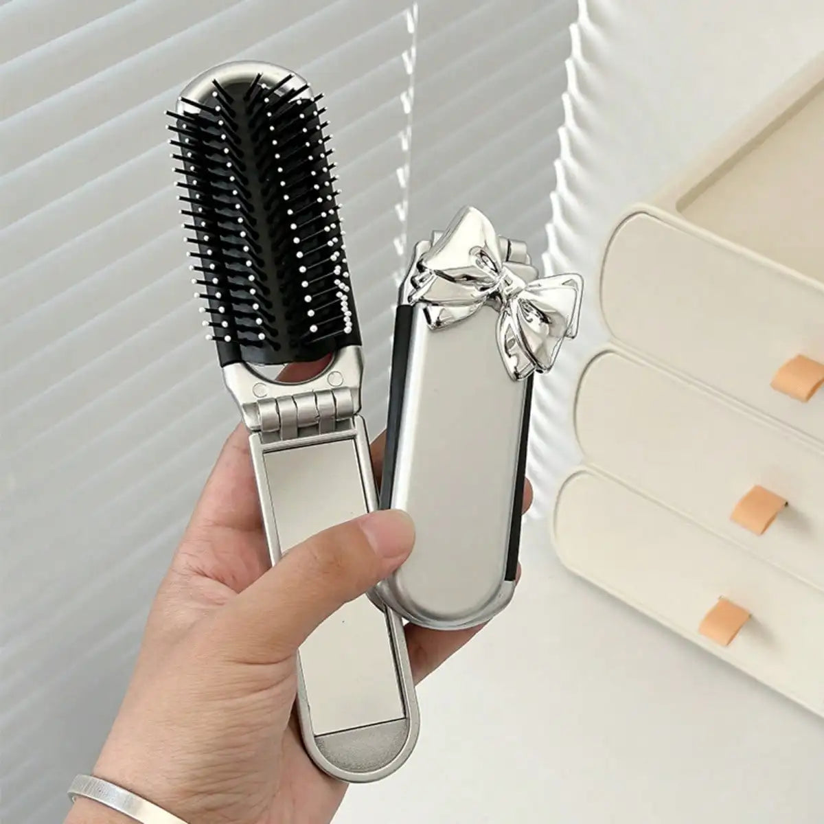 A hand holds a silver portable compact comb with an attached mirror and a black bristled comb.
