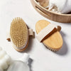 Un cepillo exfoliante corporal natural de cerdas con mango de madera y correa beige claro, junto a un cuenco de madera y una 