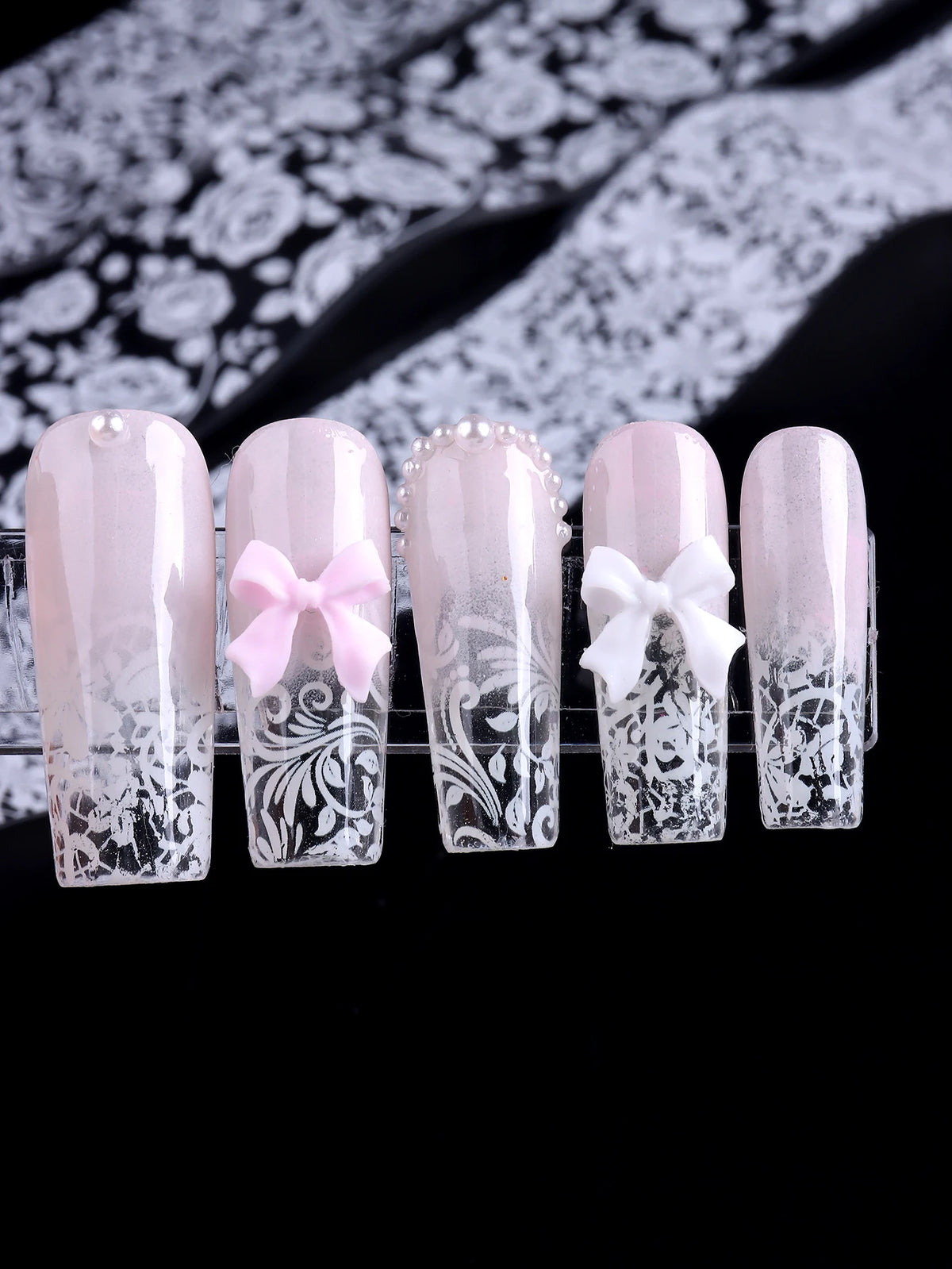 Un set de cinq autocollants pour ongles clairs et roses avec des motifs de dentelle complexes et des nœuds décoratifs.