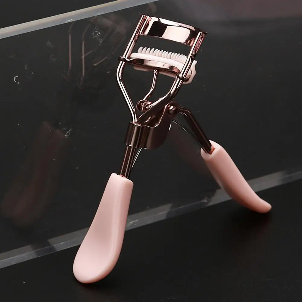 Un recourbe-cils rose professionnel avec une brosse est montré sur un fond sombre.